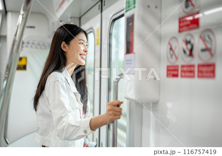 A positive Asian female is looking at the view outside while holding onto a stanchion in a sky train 116757419