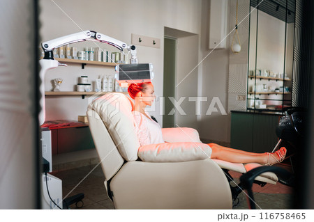 Remote wide shot of young woman client having non-invasive type of phototherapy in medical clinic during hair treatment. Blonde female sitting on chair having LED light hair photodynamic therapy. Remote wide shot of young woman client having non-invasive type of phototherapy in medical clinic during hair treatment. Blonde female sitting on chair having LED light hair photodynamic therapy. 116758465