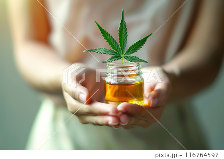 Woman Hands Presenting CBD Oil Jar with Fresh Cannabis Leaf in Soft Light Woman Hands Presenting CBD Oil Jar with Fresh Cannabis Leaf in Soft Light 116764597