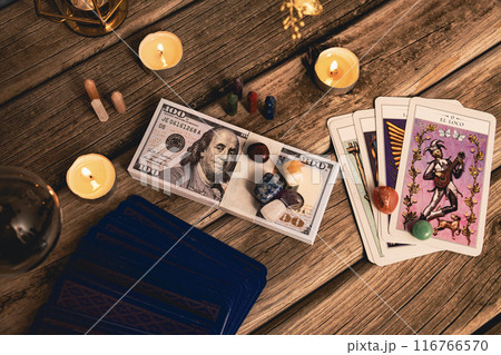 A tarot spread with The Fool card, hundred-dollar bills, and various crystals on a rustic wooden background with candles. A tarot spread with The Fool card, hundred-dollar bills, and various crystals on a rustic wooden background with candles. 116766570