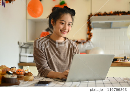 Happy young woman using laptop in a kitchen decorated for Halloween Happy young woman using laptop in a kitchen decorated for Halloween 116766868