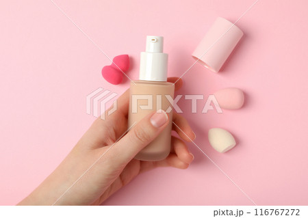 Bottle with foundation in female hand and cosmetic sponges on pink background, top view Bottle with foundation in female hand and cosmetic sponges on pink background, top view 116767272