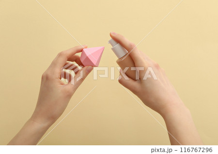 Bottle with foundation and cosmetic sponge in female hands on beige background 116767296