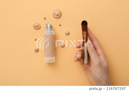 Cosmetic brush in female hand, bottle and drops of foundation on beige background, top view 116767297