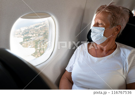 An elderly woman travels. Flying in a plane wearing a mask. Passenger. Watch the porthole. Look at the ground from above. Active lifestyle. An elderly woman travels. Flying in a plane wearing a mask. Passenger. Watch the porthole. Look at the ground from above. Active lifestyle. 116767536