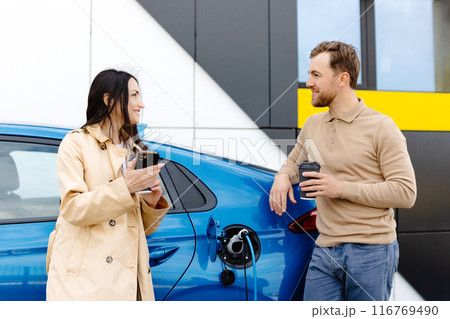 Young couple man and woman traveling by electric car having stop at charging station. Boyfriend plugging in cable to charge. Man talking with girlfriend, holding cup drinking hot coffee smiling Young couple man and woman traveling by electric car having stop at charging station. Boyfriend plugging in cable to charge. Man talking with girlfriend, holding cup drinking hot coffee smiling 116769490
