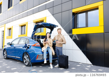 Young friends charging automobile battery from public station and standing near parking for electric car. Man and woman talking and drinking coffee in city Young friends charging automobile battery from public station and standing near parking for electric car. Man and woman talking and drinking coffee in city 116769493