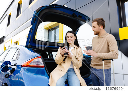 Young friends charging automobile battery from public station and standing near parking for electric car. Man and woman talking and drinking coffee in city 116769494
