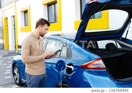 A young man is charging an electric car while standing at a station and holding a tablet 116769495