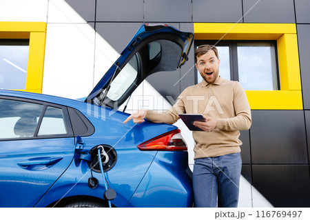 A young man is charging an electric car while standing at a station and holding a tablet 116769497