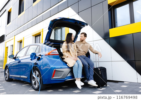 Young couple man and woman traveling by electric car having stop at charging station. Boyfriend and girlfriend sitting in the car trunk and smiling 116769498