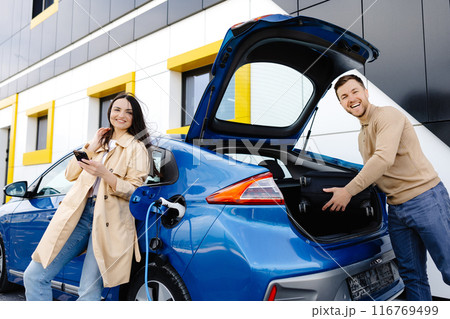 Young couple man and woman traveling by electric car having stop at charging station. The man taking a suitcase from the trunk. Man talking with girlfriend and she holding mobile phone in hand Young couple man and woman traveling by electric car having stop at charging station. The man taking a suitcase from the trunk. Man talking with girlfriend and she holding mobile phone in hand 116769499