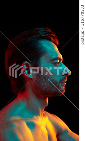 Profile view portrait of handsome man posing shirtless, showing well kept skin and body against black studio background in neon light 116770533
