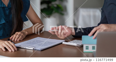 Real Estate Agent Discussing Contract with Client at Office Desk with Model House and Calculator Real Estate Agent Discussing Contract with Client at Office Desk with Model House and Calculator 116771499