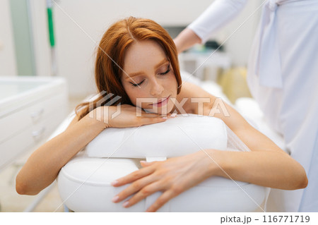 Closeup portrait of calm woman getting moxibustion treatment on back lying on massage table with closed eyes. Female client lying on spa bed during moxa smoke therapy. Concept of alternative medicine. Closeup portrait of calm woman getting moxibustion treatment on back lying on massage table with closed eyes. Female client lying on spa bed during moxa smoke therapy. Concept of alternative medicine. 116771719