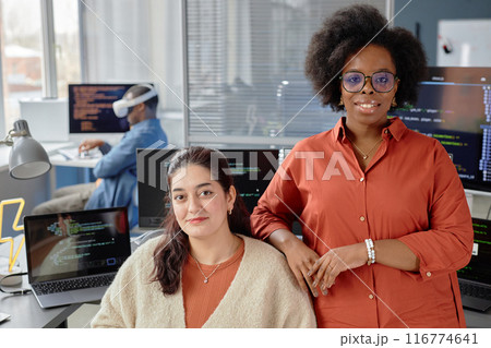 Portrait shot of two female multiethnic VR developers at working desk with computers looking at camera and smiling in IT company office Portrait shot of two female multiethnic VR developers at working desk with computers looking at camera and smiling in IT company office 116774641