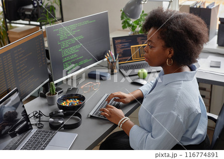 High-angle side view shot of African American female programmer working on computer reading multiple screens with code lines running High-angle side view shot of African American female programmer working on computer reading multiple screens with code lines running 116774891