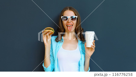 Happy cheerful young woman eating burger fast food holding cup of coffee, teenage girl having fun Happy cheerful young woman eating burger fast food holding cup of coffee, teenage girl having fun 116776258