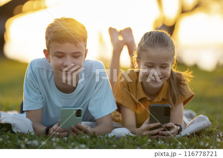 Happy teenagers girl and boy messaging on their cellphones outdoors in summer park Happy teenagers girl and boy messaging on their cellphones outdoors in summer park 116778721