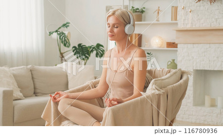 Audio meditation. Peaceful balance. Wellness leisure. Relaxed woman in headphones sitting in comfortable position in armchair listening to mantra at home. 116780936