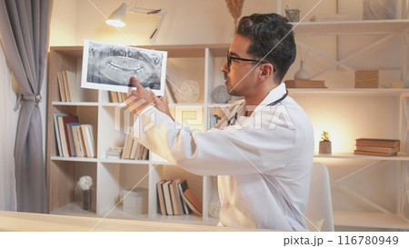Dentist consultation. Teeth radiography. Video appointment. Professional man with x ray of mouth explaining diagnosis at online meeting sitting in clinic office workplace interior. 116780949