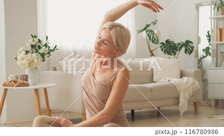 Morning practice. Sportive lifestyle. Healthy habit. Calm woman sitting yoga position on floor stretching back side hand up in light home interior. Morning practice. Sportive lifestyle. Healthy habit. Calm woman sitting yoga position on floor stretching back side hand up in light home interior. 116780986