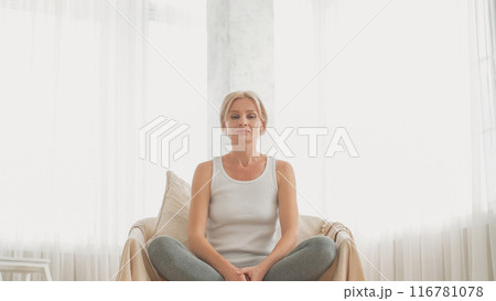 Relaxation practice. Peaceful morning. Home meditation. Harmony smiling woman sitting in lotus pose on couch breathing in light interior copy space. 116781078
