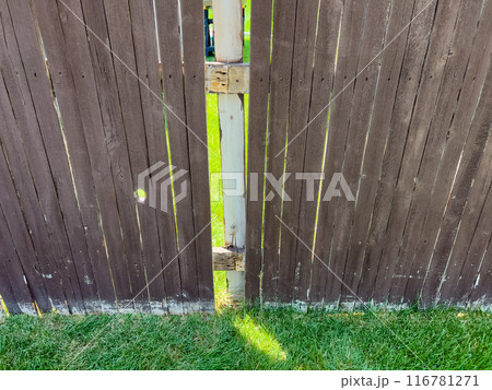 A backyard wooden fence showing a small gap between its panels. The brown fence, weathered with time, is highlighted by the sunlit grass visible through the gap. This image captures the rustic charm 116781271