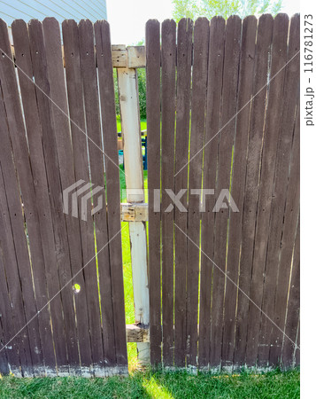 A backyard wooden fence showing a small gap between its panels. The brown fence, weathered with time, is highlighted by the sunlit grass visible through the gap. This image captures the rustic charm 116781273