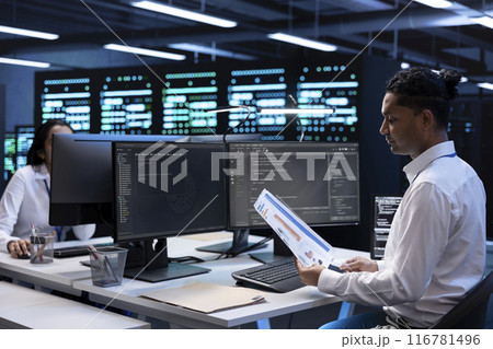 Engineer in data center doing yearly maintenance checkup to prolong hardware life span, looking over documents, monitoring server infrastructure and reading specifications paperwork 116781496