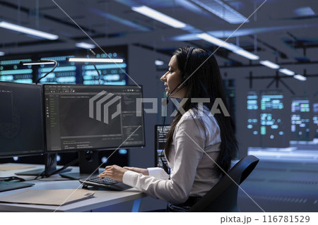 Data center support expert supporting engineering team doing equipment maintenance, talking in headset microphone. Woman in server farm coordinating technicians using mic Data center support expert supporting engineering team doing equipment maintenance, talking in headset microphone. Woman in server farm coordinating technicians using mic 116781529