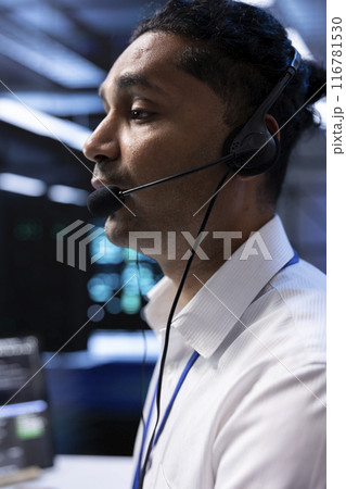 Data center agent supporting engineering team doing hardware maintenance, talking in headset microphone. Worker in high tech server room facility coordinating technicians using mic Data center agent supporting engineering team doing hardware maintenance, talking in headset microphone. Worker in high tech server room facility coordinating technicians using mic 116781530