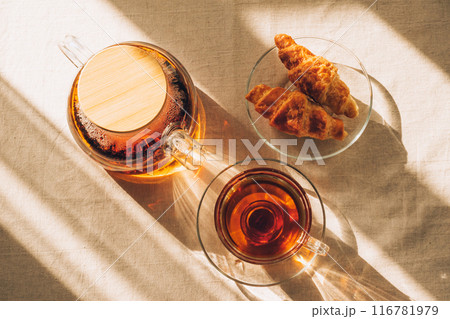Top view of glass teapot with brewed tea, cup of tea and croissants on table under sunlight casting shadows 116781979