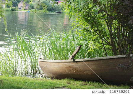 Old wooden boat with oars on lake shore. Houses and buildings. Water transportation and nature in summer. Old wooden boat with oars on lake shore. Houses and buildings. Water transportation and nature in summer. 116786201