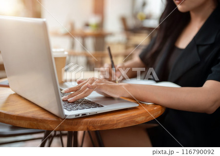 A businesswoman working remotely from a cafe, typing on the laptop keyboard and taking note. A businesswoman working remotely from a cafe, typing on the laptop keyboard and taking note. 116790092