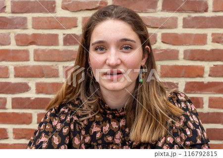 A stylish young woman with long hair in a patterned blouse poses against a brick wall 116792815