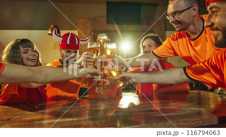 Fans in matching orange shirts and fun hats cheerfully toast with their beers at bar, celebrating with wide smiles. 116794063