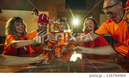 Friends, football fans in orange shirts raise their glasses in celebratory toast, their faces beaming with happiness and energy. Friends, football fans in orange shirts raise their glasses in celebratory toast, their faces beaming with happiness and energy. 116794064