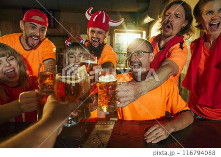 Enthusiastic fans in orange shirts, some wearing hats, cheer and clink glasses of beer at bar, their faces full of excitement and joy. 116794088