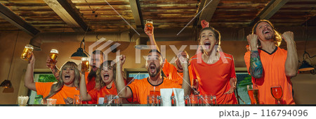 Banner. At a bar, excited people in orange shirts celebrate with raised glasses victory of favorite sport team while watching its match on TV. 116794096