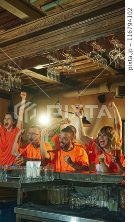 Excited young men and women in orange shirts celebrate at bar, raising their beers and cheering, victory of favorite sport team. 116794102