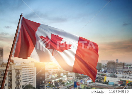 A red and white Canadian flag is flying high in the sky over a city 116794959