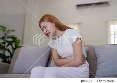Young Woman Experiencing Stomachache While Sitting on Couch in Living Room, Expressing Pain and Discomfort Young Woman Experiencing Stomachache While Sitting on Couch in Living Room, Expressing Pain and Discomfort 116797080
