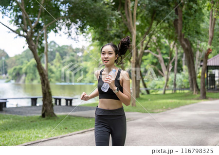 Young Woman Jogging in Park with Water Bottle, Enjoying Outdoor Exercise and Fresh Air Young Woman Jogging in Park with Water Bottle, Enjoying Outdoor Exercise and Fresh Air 116798325