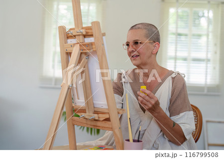Elderly Woman Painting on Canvas in Bright Studio with Natural Light and Wooden Easel 116799508