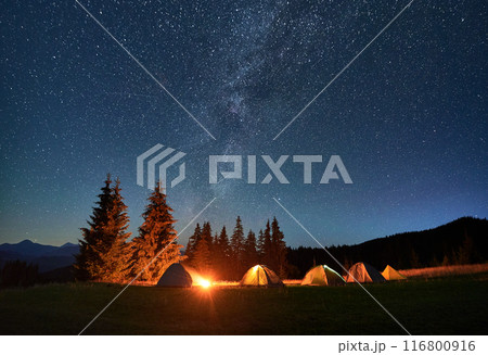 Night camping in mountains under starry sky. Tourist tents in campsite near burning campfire under beautiful sky full of stars with Milky way above forest. Concept of tourism and traveling. 116800916