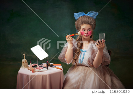 Blushing cheeks. Portrait of young woman, princess sitting at table and making bright makeup against vintage green background Blushing cheeks. Portrait of young woman, princess sitting at table and making bright makeup against vintage green background 116801020