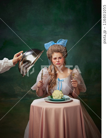 Woman in historical gown with blue bow eating cauliflower with pretty face against vintage green background. Following healthy eating 116801055