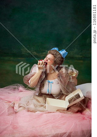 Sweet tooth. Young woman in period attire emotional eating dessert in bed against vintage green background. Delicious baked goods 116801181