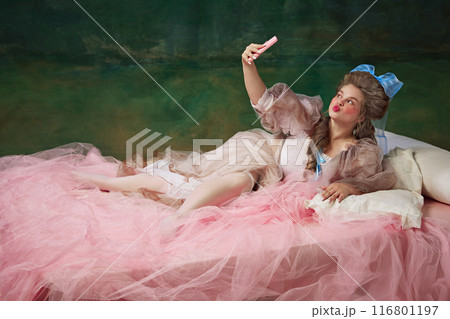 Elegant woman, royal person lying on bed with pink fabric and taking selfie with mobile phone against vintage green background Elegant woman, royal person lying on bed with pink fabric and taking selfie with mobile phone against vintage green background 116801197